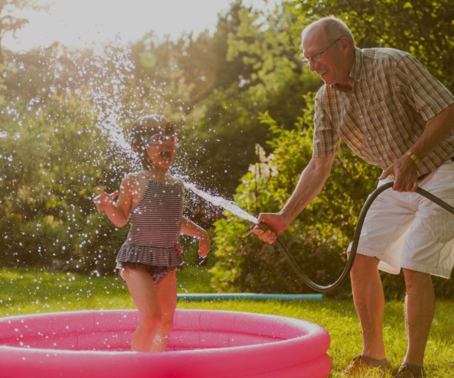 A grandfather and his granddaughter are playing in the year with water. He is spraying her with a hose and she's laughing.