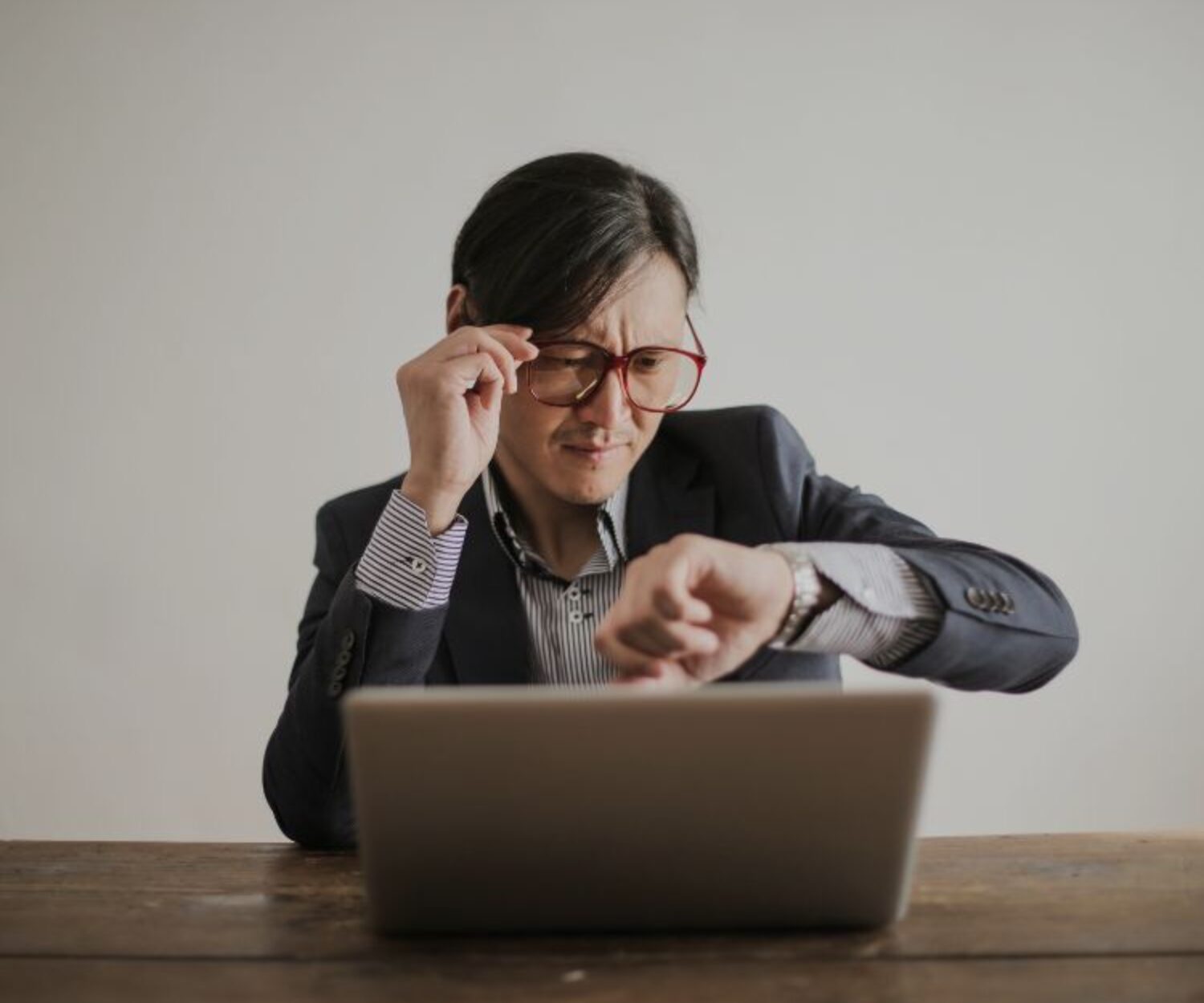 Asian male sitting in front of the a laptop looking at his watch with a concerned look on his face